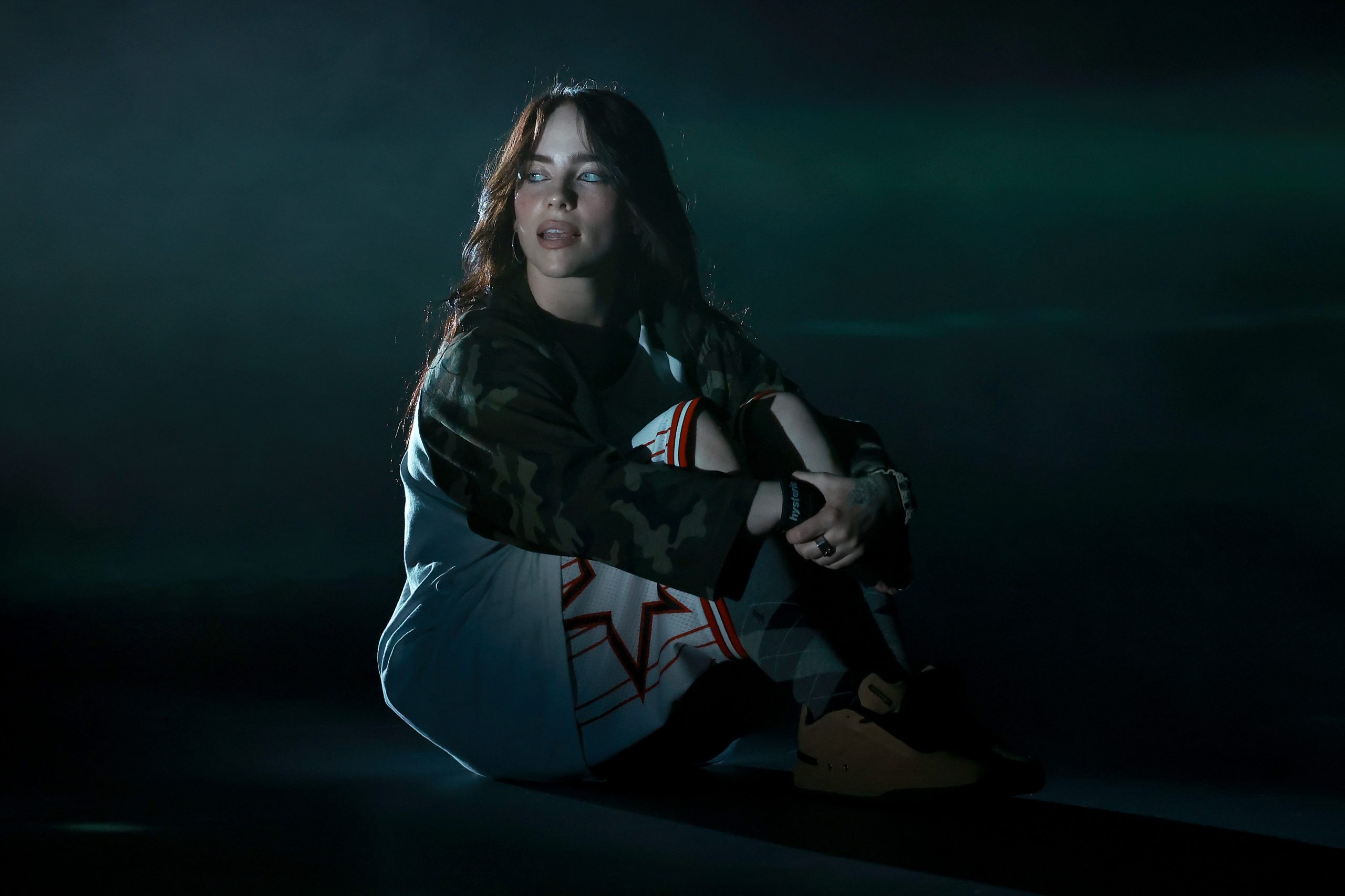 billie eilish sits on the ground in a dark room with her arms wrapped around bent knees. She stares off camera.