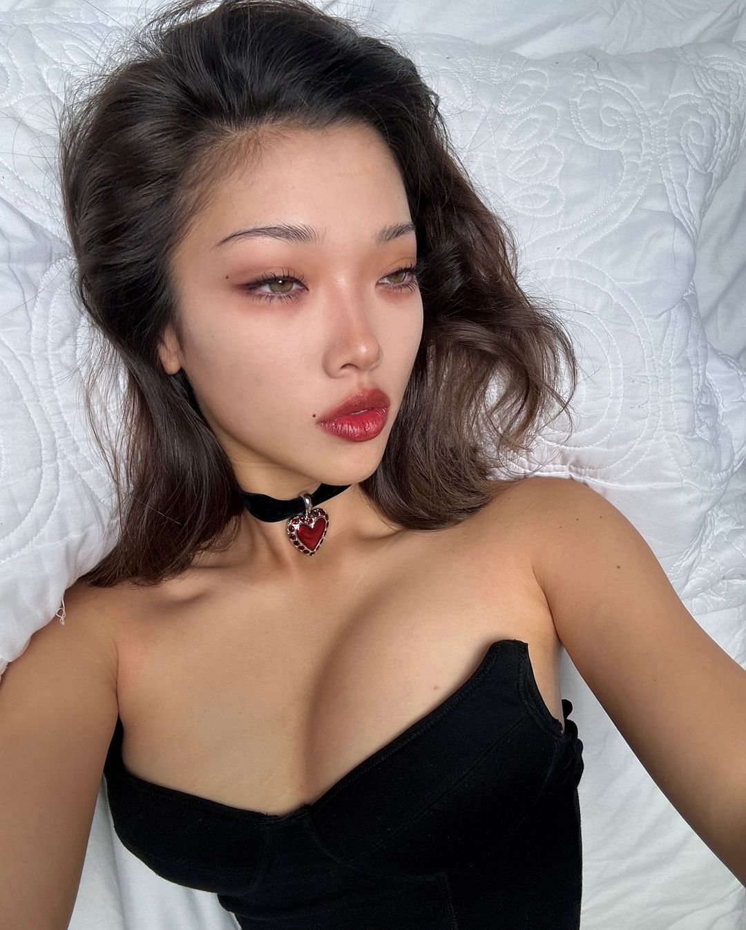 the rumoured girlfriend of nicholas galitzine, cameron valentina eyre, lies on a bed, taking a selfie from above. She wears a black corset top and black choker, and her dark hair spills across the bed [nicholas galitzine girlfriend]